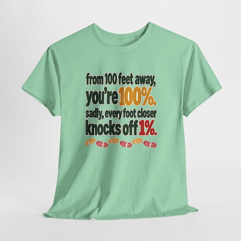 May include: A light green short-sleeve t-shirt featuring the black text: from 100 feet away, you're 100%. sadly, every foot closer knocks off 1%. The text includes orange and red accents, with small orange and red shoe print icons below.