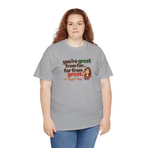 May include: A heather gray short-sleeve t-shirt featuring a graphic print with the text "you're great from far... far from great." The design includes a cartoon illustration of a person with brown hair and several small ticket stubs.
