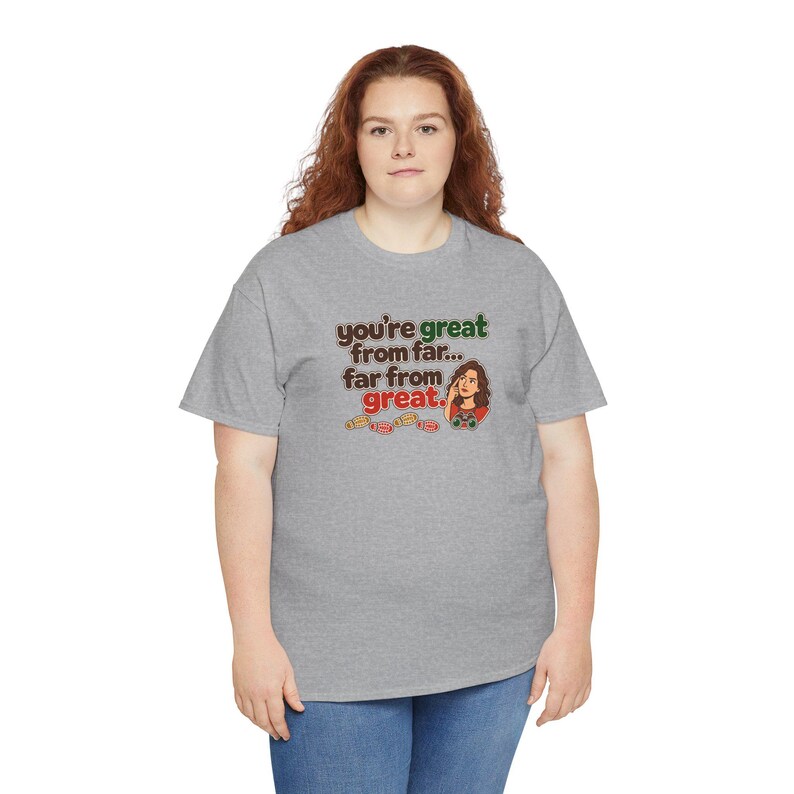 May include: A heather gray short-sleeve t-shirt featuring a graphic print with the text "you're great from far... far from great." The design includes a cartoon illustration of a person with brown hair and several small ticket stubs.