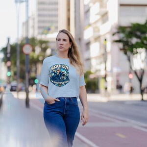 May include: A light blue short-sleeve t-shirt featuring a circular graphic of a pilot wearing sunglasses and headphones with a small robot. The text reads WATCH OUT! I'M ON AUTOPILOT NOW with a small airplane icon at the bottom.