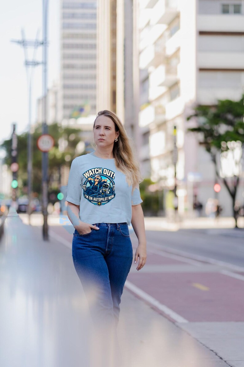 May include: A light blue short-sleeve t-shirt featuring a circular graphic of a pilot wearing sunglasses and headphones with a small robot. The text reads WATCH OUT! I'M ON AUTOPILOT NOW with a small airplane icon at the bottom.