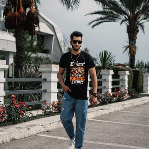 Puede incluir: Una camiseta negra de manga corta con un estampado gr&aacute;fico de un pu&ntilde;o y la ecuaci&oacute;n f&iacute;sica E=1/2mv2 con el texto AVOID MY KINETIC ENERGY en naranja y blanco, combinada con vaqueros de mezclilla azul claro desgastados y zapatillas blancas.