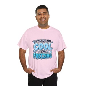 May include: A light pink short-sleeved t-shirt featuring a graphic print with the text "YOU'RE SO COOL, I'M FREEZING." The lettering is styled with an icy, blue-toned texture, surrounded by small snowflake and ice cube illustrations.