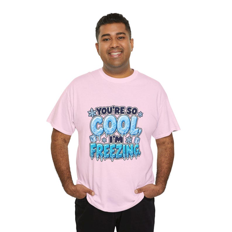 May include: A light pink short-sleeved t-shirt featuring a graphic print with the text "YOU'RE SO COOL, I'M FREEZING." The lettering is styled with an icy, blue-toned texture, surrounded by small snowflake and ice cube illustrations.