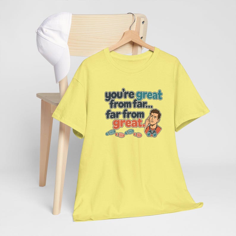 May include: A light yellow short-sleeve t-shirt featuring a graphic of a person with binoculars and the text "you're great from far... far from great." The design includes small colorful footprints below the text and a white baseball cap.