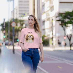 May include: A light pink short-sleeved t-shirt featuring a graphic illustration of a Shih Tzu dog on the chest, with the text Shih Tzu printed in black above the dog's head. The shirt has a crew neckline and a straight hemline.