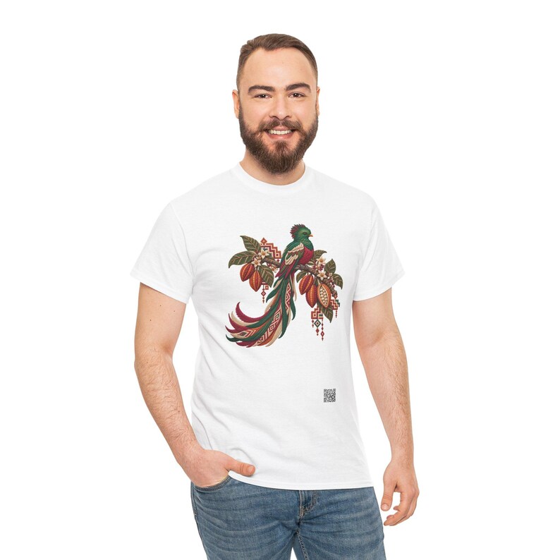 May include: A white t-shirt featuring a detailed graphic of a quetzal bird perched on a branch with cacao pods. The bird has vibrant green, red, and white plumage with a long tail. The design includes geometric patterns and small flowers.