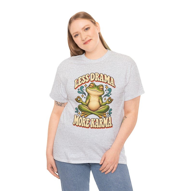 May include: A light gray t-shirt featuring a graphic of a green frog sitting in a lotus position with eyes closed. The text above the frog reads LESS DRAMA and the text below reads MORE KARMA in a bold, retro-style font with blue accents.