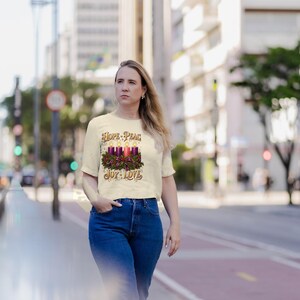 May include: A cream-colored short-sleeve t-shirt featuring a graphic of four lit candles surrounded by a green wreath and a red bow. The text "HOPE - PEACE" is printed above the candles, and "JOY - LOVE" is printed below the graphic.