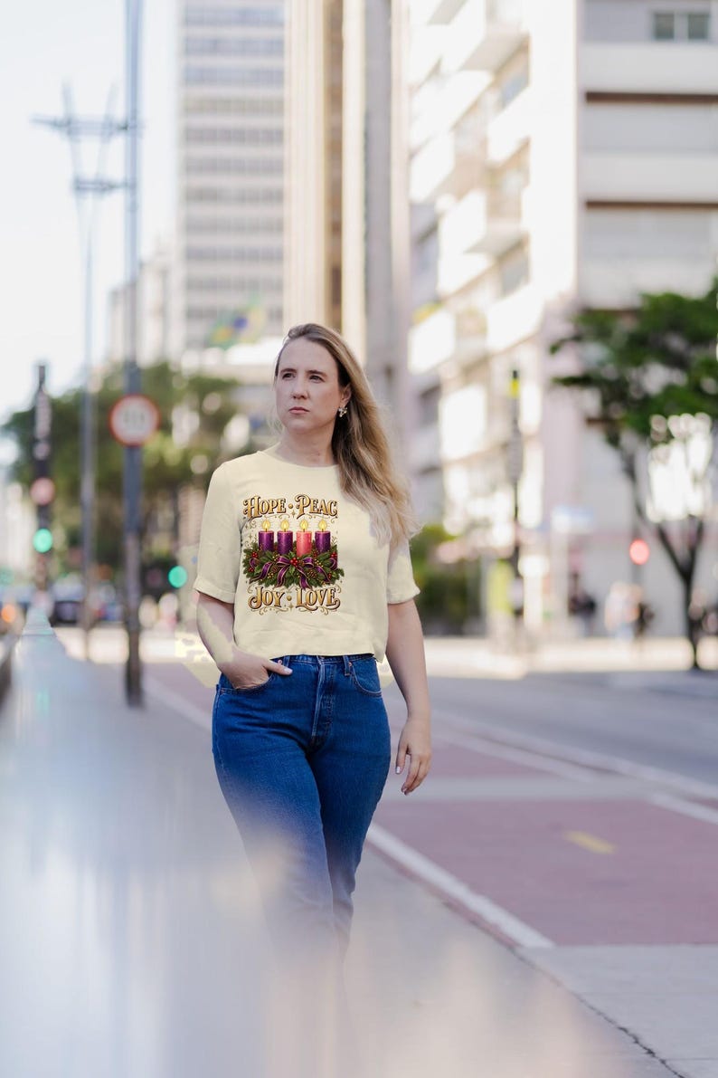 May include: A cream-colored short-sleeve t-shirt featuring a graphic of four lit candles surrounded by a green wreath and a red bow. The text "HOPE - PEACE" is printed above the candles, and "JOY - LOVE" is printed below the graphic.