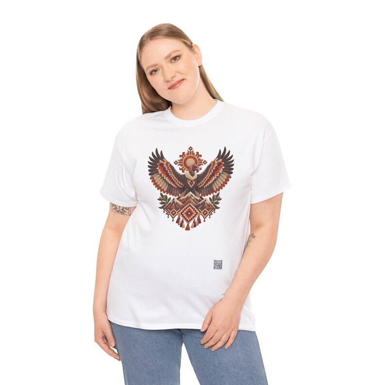 May include: A white short-sleeve t-shirt featuring a central graphic of a stylized eagle with spread wings, rendered in shades of brown, red, and orange with geometric patterns. A small square QR code is printed near the bottom right hem.