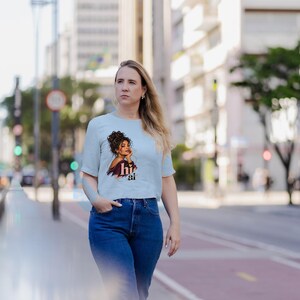 May include: A light blue short-sleeved t-shirt featuring a graphic print of a person with dark curly hair resting their chin on their hand, accompanied by the text "hi ai" in a stylized brown font, paired with dark blue denim jeans.