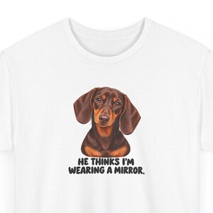 May include: A white t-shirt featuring a brown and black dachshund illustration centered on the chest. Below the dog, the text reads: HE THINKS I'M WEARING A MIRROR. The shirt has a crew neckline and short sleeves, providing a clean, simple look.