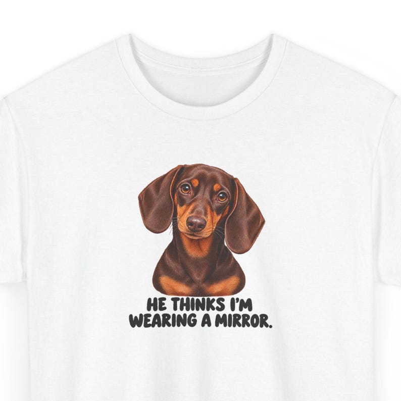 May include: A white t-shirt featuring a brown and black dachshund illustration centered on the chest. Below the dog, the text reads: HE THINKS I'M WEARING A MIRROR. The shirt has a crew neckline and short sleeves, providing a clean, simple look.