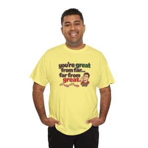 May include: A light yellow short-sleeve t-shirt featuring the text "you're great from far... far from great." in brown and green lettering, accompanied by a cartoon illustration of a person holding binoculars and small footprints.