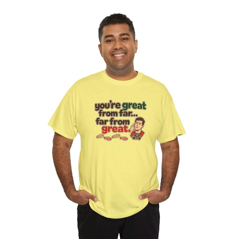 May include: A light yellow short-sleeve t-shirt featuring the text "you're great from far... far from great." in brown and green lettering, accompanied by a cartoon illustration of a person holding binoculars and small footprints.