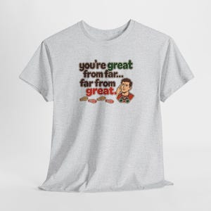 May include: A light gray short-sleeve t-shirt featuring a graphic of a person looking through binoculars with the text: you're great from far... far from great. Small footprints are printed below the text in brown, orange, and red colors.