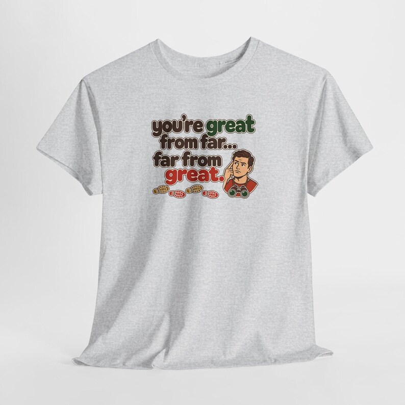 May include: A light gray short-sleeve t-shirt featuring a graphic of a person looking through binoculars with the text: you're great from far... far from great. Small footprints are printed below the text in brown, orange, and red colors.