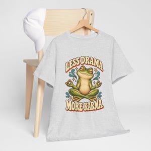 May include: A light gray short-sleeve t-shirt featuring a graphic of a green frog sitting in a lotus position with eyes closed. The text above and below the frog reads "LESS DRAMA" and "MORE KARMA" in a bold, curved, orange-outlined font.