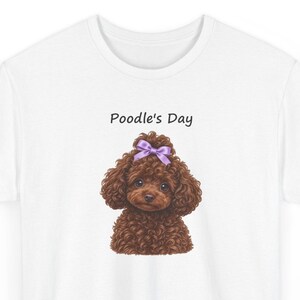 May include: A white t-shirt featuring a graphic of a brown curly-haired poodle with a purple bow on its head. Above the dog, the text Poodle's Day is printed in a simple black font. The design is centered on the chest of the shirt.