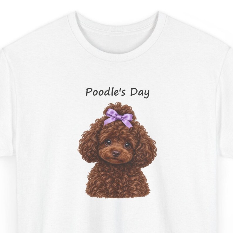 May include: A white t-shirt featuring a graphic of a brown curly-haired poodle with a purple bow on its head. Above the dog, the text Poodle's Day is printed in a simple black font. The design is centered on the chest of the shirt.