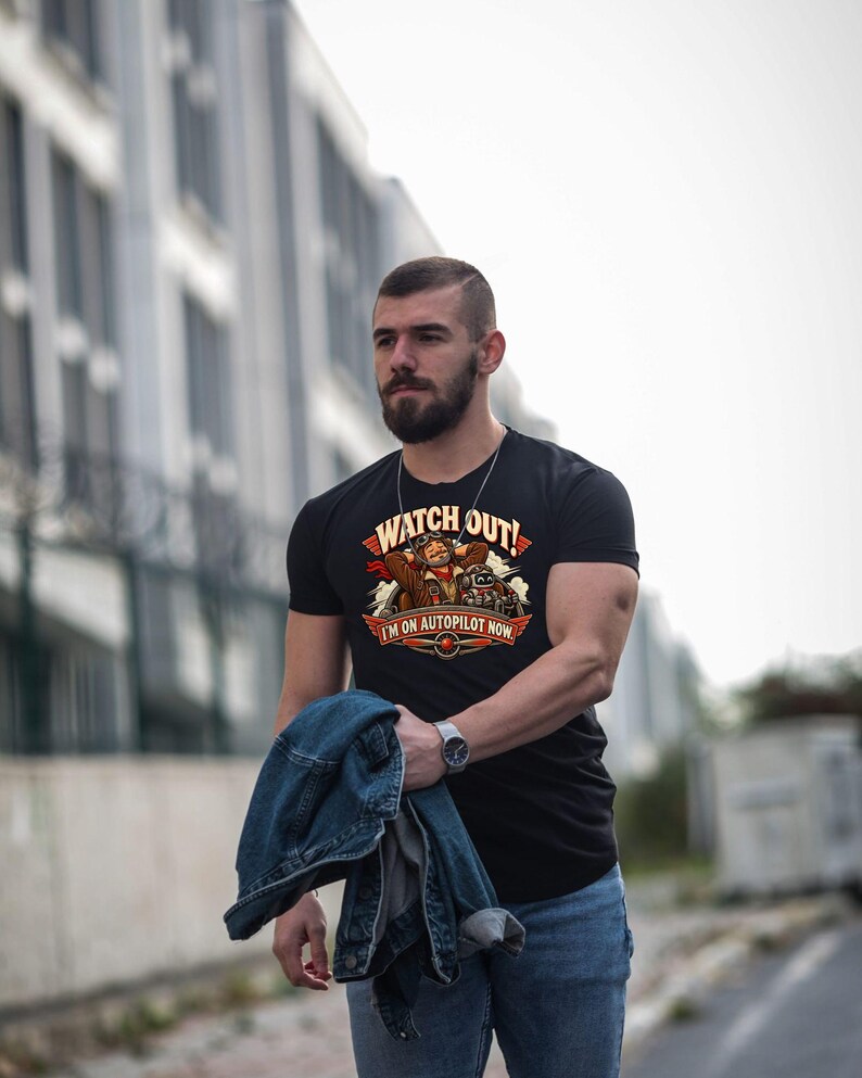 May include: A black short-sleeve t-shirt featuring a graphic of a pilot with goggles and a robot in a cockpit. The text reads WATCH OUT! I'M ON AUTOPILOT NOW. The design includes red, orange, and brown accents with a propeller icon at the bottom.