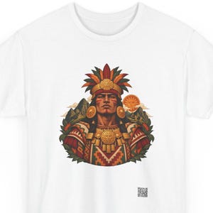 May include: A white t-shirt featuring a graphic illustration of a person wearing a feathered headdress and gold jewelry. The design includes a sun, mountain peaks, and geometric patterns in red, orange, and green. A small QR code is printed at the bottom.