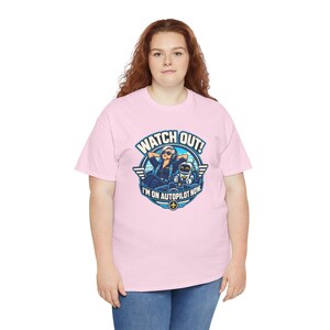 May include: A light pink short-sleeve t-shirt featuring a circular graphic of a pilot and a robot in a cockpit. The text reads WATCH OUT! I'M ON AUTOPILOT NOW. The design includes blue and white colors with wings and an airplane icon.