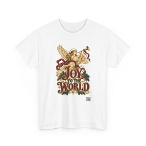 May include: A white t-shirt featuring a graphic of an angel with wings playing a golden trumpet. Below the angel, the text reads JOY TO THE WORLD in stylized red and gold lettering, surrounded by green holly leaves and small gold stars.