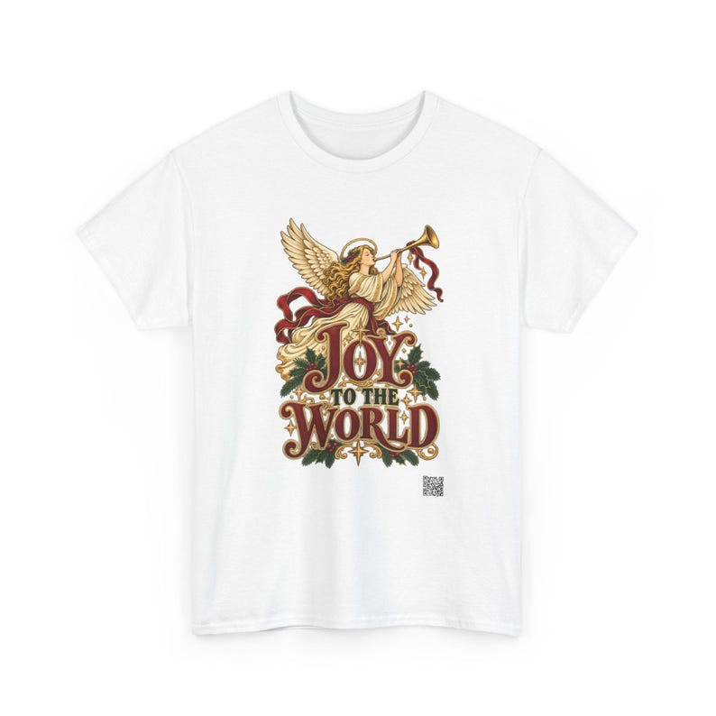 May include: A white t-shirt featuring a graphic of an angel with wings playing a golden trumpet. Below the angel, the text reads JOY TO THE WORLD in stylized red and gold lettering, surrounded by green holly leaves and small gold stars.