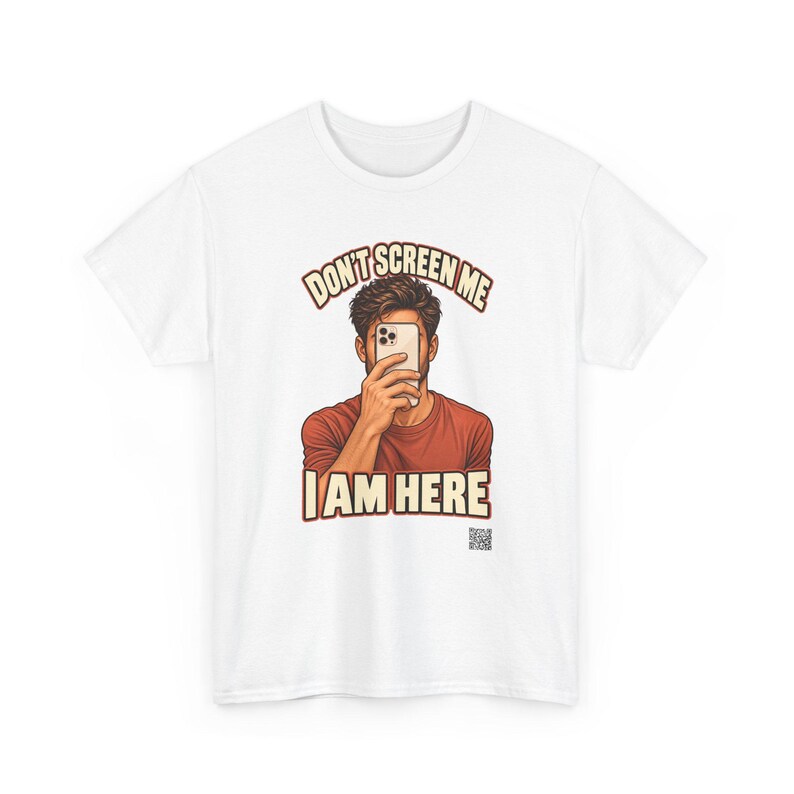 Op de afbeelding: Een wit t-shirt met een afbeelding van een persoon in een roestkleurig shirt die een smartphone voor het gezicht houdt. De tekst erboven luidt DON'T SCREEN ME en de tekst eronder I AM HERE, met een kleine QR-code rechtsonder.