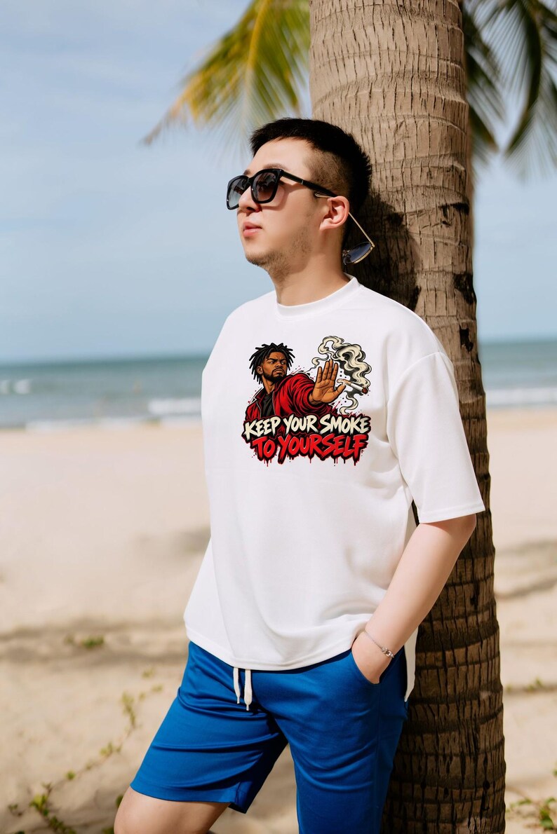 May include: A white short-sleeved t-shirt featuring a graphic print of a person in a red jacket holding a hand out with smoke rising from a cigarette. The text "KEEP YOUR SMOKE TO YOURSELF" is printed in bold black and red lettering below the graphic.