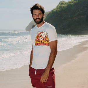 May include: A white short-sleeved t-shirt featuring a graphic of a cigarette inside a red prohibition circle surrounded by orange flowers and smoke, with the text "YOUR SMOKE IS NOT MY VIBE" in a retro orange and yellow font.