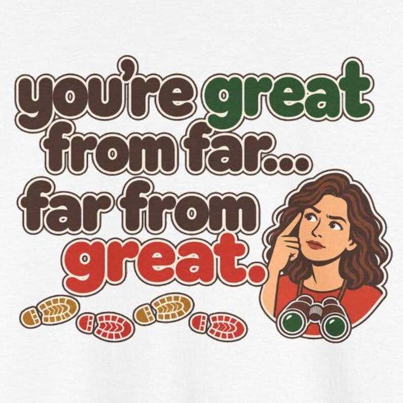 May include: A white t-shirt featuring the brown and green text "you're great from far... far from great." with a cartoon illustration of a person with brown hair wearing binoculars and red shirt, accompanied by brown and red shoe prints.