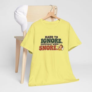 May include: A light yellow short-sleeve t-shirt featuring the text HARD TO IGNORE. ESPECIALLY WHEN I SNORE. in green and red lettering, accompanied by a cartoon illustration of a person sleeping and snoring on a white pillow.