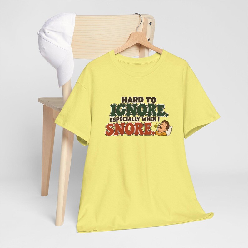 May include: A light yellow short-sleeve t-shirt featuring the text HARD TO IGNORE. ESPECIALLY WHEN I SNORE. in green and red lettering, accompanied by a cartoon illustration of a person sleeping and snoring on a white pillow.