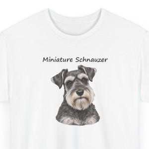 May include: A white t-shirt featuring a detailed illustration of a Miniature Schnauzer dog with black and grey fur and a light-colored beard. The text "Miniature Schnauzer" is printed in a simple black font above the dog's head.