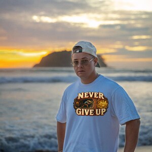 Puede incluir: Una camiseta blanca de manga corta con un estampado gr&aacute;fico de un minero trabajando en una cueva y el texto NEVER GIVE UP en letras may&uacute;sculas de color beige. El dise&ntilde;o incluye una peque&ntilde;a ilustraci&oacute;n de una persona extrayendo oro.