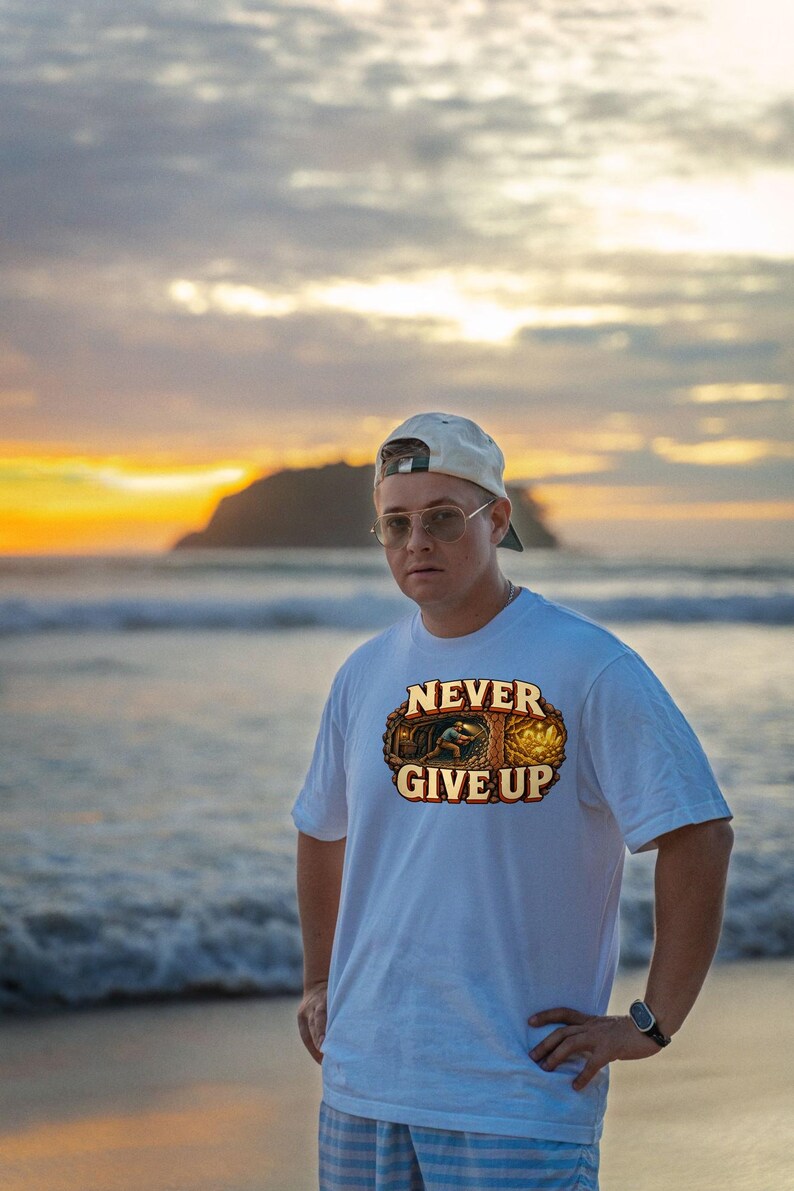 Puede incluir: Una camiseta blanca de manga corta con un estampado gr&aacute;fico de un minero trabajando en una cueva y el texto NEVER GIVE UP en letras may&uacute;sculas de color beige. El dise&ntilde;o incluye una peque&ntilde;a ilustraci&oacute;n de una persona extrayendo oro.