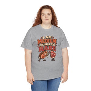 May include: A heather gray short-sleeve t-shirt featuring a graphic of two cartoon steak pieces. The text reads I AM MEDIUM LOOKING FOR RARE. One steak holds a magnifying glass, and a small red heart is positioned between the two steak characters.