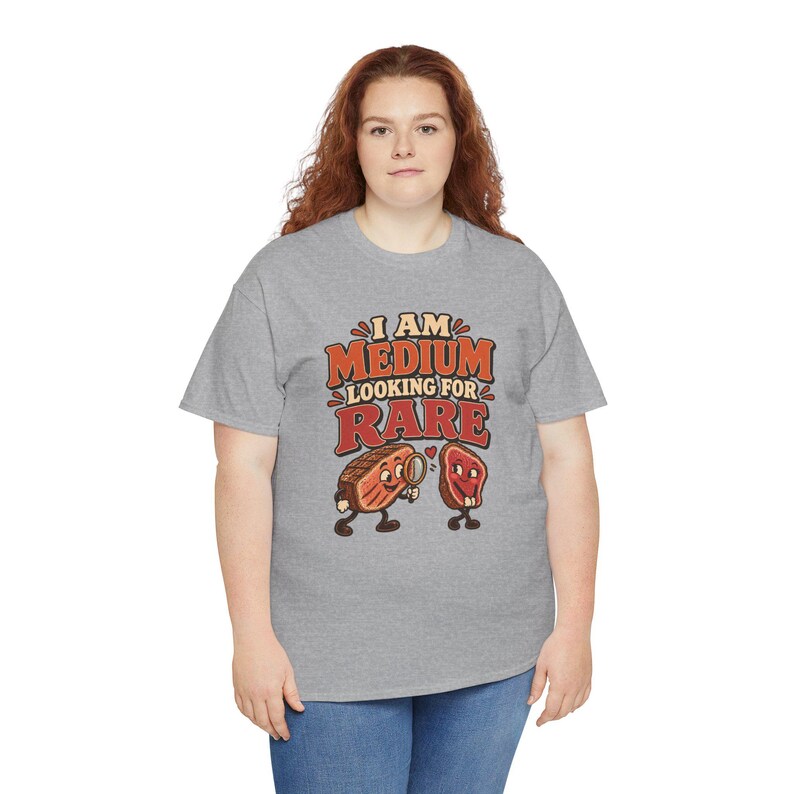 May include: A heather gray short-sleeve t-shirt featuring a graphic of two cartoon steak pieces. The text reads I AM MEDIUM LOOKING FOR RARE. One steak holds a magnifying glass, and a small red heart is positioned between the two steak characters.