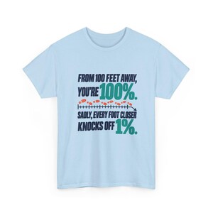May include: A light blue t-shirt featuring dark navy and teal text that reads: FROM 100 FEET AWAY, YOU'RE 100%. SADLY, EVERY FOOT CLOSER KNOCKS OFF 1%. The design includes a horizontal line with small orange footprints above it.