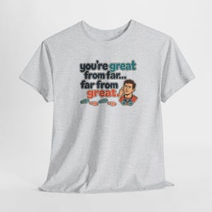 May include: A light gray short-sleeved t-shirt featuring the text "you're great from far... far from great." alongside an illustration of a person looking through binoculars and four small colorful footprints below the text.