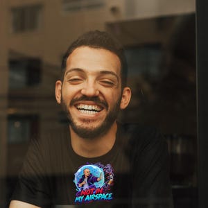 Może przedstawiać: Czarny t-shirt z nadrukiem graficznym przedstawiającym osobę w niebieskiej bańce energetycznej z napisem NOT IN MY AIRSPACE czerwonymi i niebieskimi literami. Wz&oacute;r zawiera efekty fioletowego dymu i stylizowany gest dłoni.