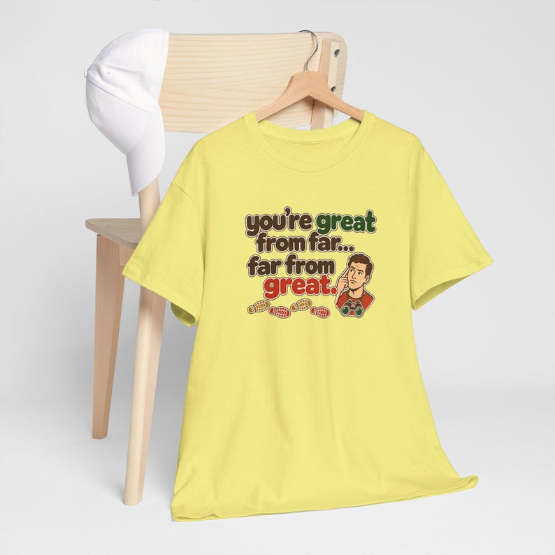 May include: A light yellow short-sleeve t-shirt featuring the text "you're great from far... far from great." and an illustration of a person looking through binoculars, accompanied by a white baseball cap hanging on a wooden chair.