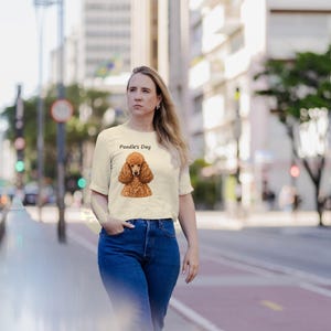 May include: A cream-colored short-sleeve t-shirt featuring a graphic illustration of a brown poodle and the text Poodle's Day printed in black above the dog. The shirt is paired with blue denim jeans, creating a casual outfit.