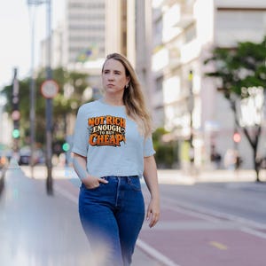May include: A light blue short-sleeved t-shirt featuring the orange and black text "NOT RICH ENOUGH TO BUY CHEAP" printed on the chest, paired with dark blue denim jeans. The shirt has a crew neckline and a standard fit.
