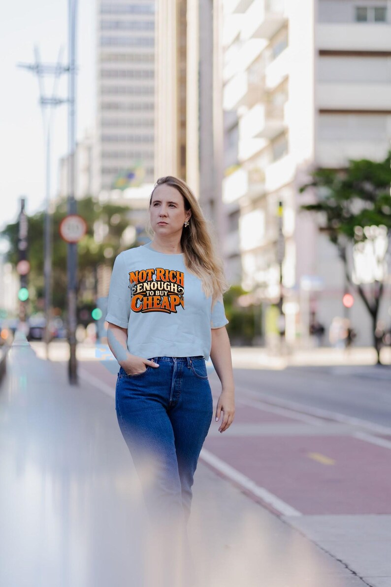 May include: A light blue short-sleeved t-shirt featuring the orange and black text "NOT RICH ENOUGH TO BUY CHEAP" printed on the chest, paired with dark blue denim jeans. The shirt has a crew neckline and a standard fit.