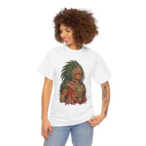 May include: A white short-sleeved t-shirt featuring a graphic print of an Aztec warrior wearing a large green feathered headdress, a gold sun medallion, and a red and green patterned cape. The design is centered on the chest of the shirt.