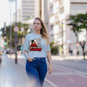 May include: A light blue short-sleeve t-shirt featuring a graphic illustration of a person in a red hoodie holding a cigarette with smoke swirling around. The text "KEEP YOUR SMOKE TO YOURSELF" is printed in bold red and black lettering below the figure.